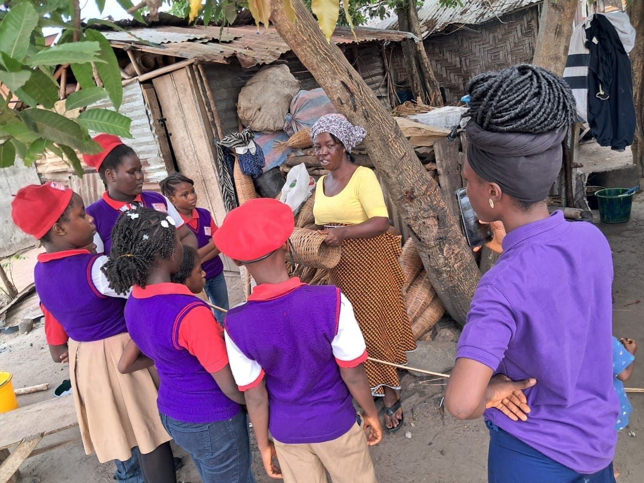 Children in purple shirts and red berets gathered around a woman showing them how to weave a fishing basket.