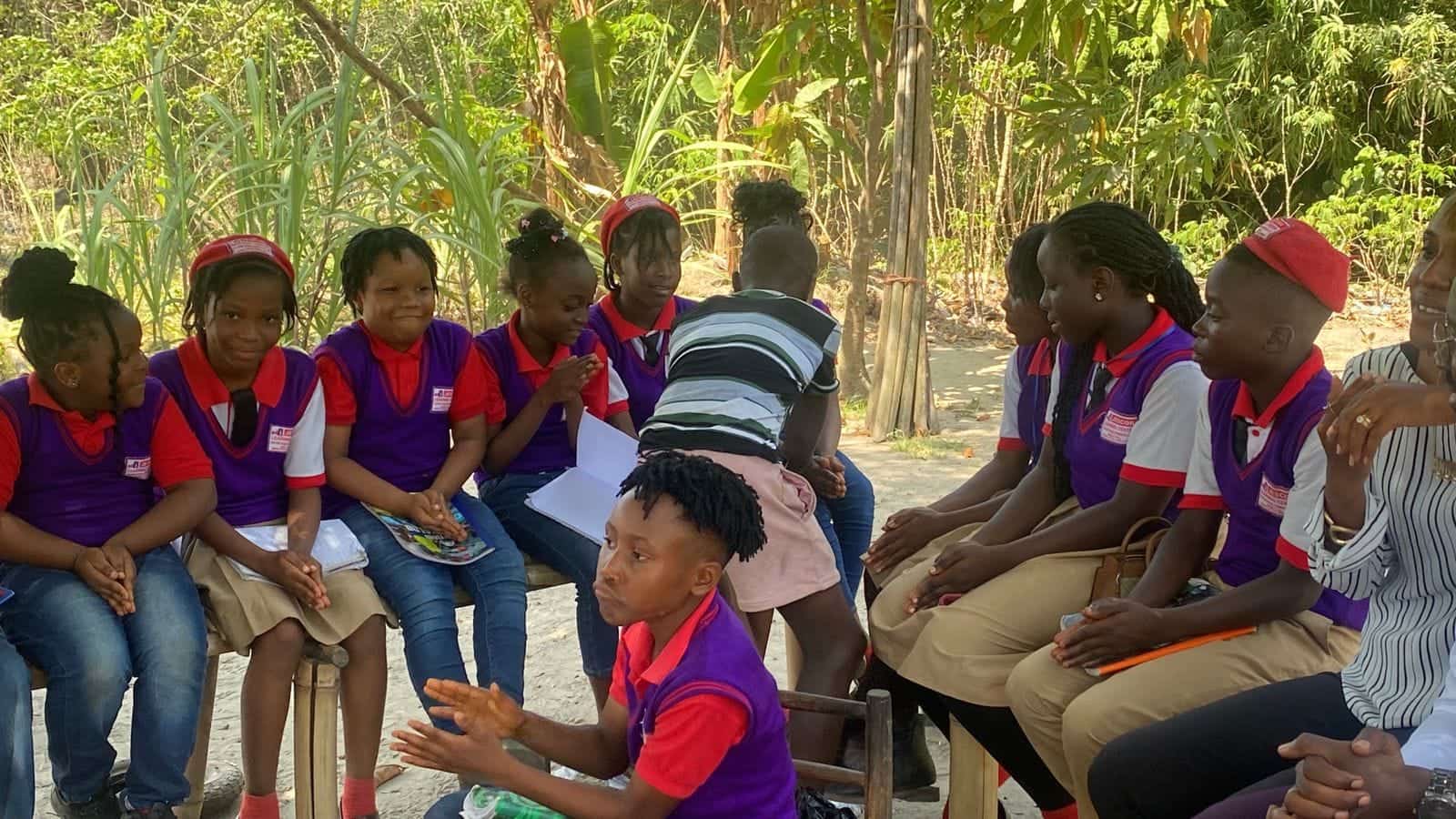Children in purple and red school uniforms sitting on benches in a circle outdoors while one girl speaks.