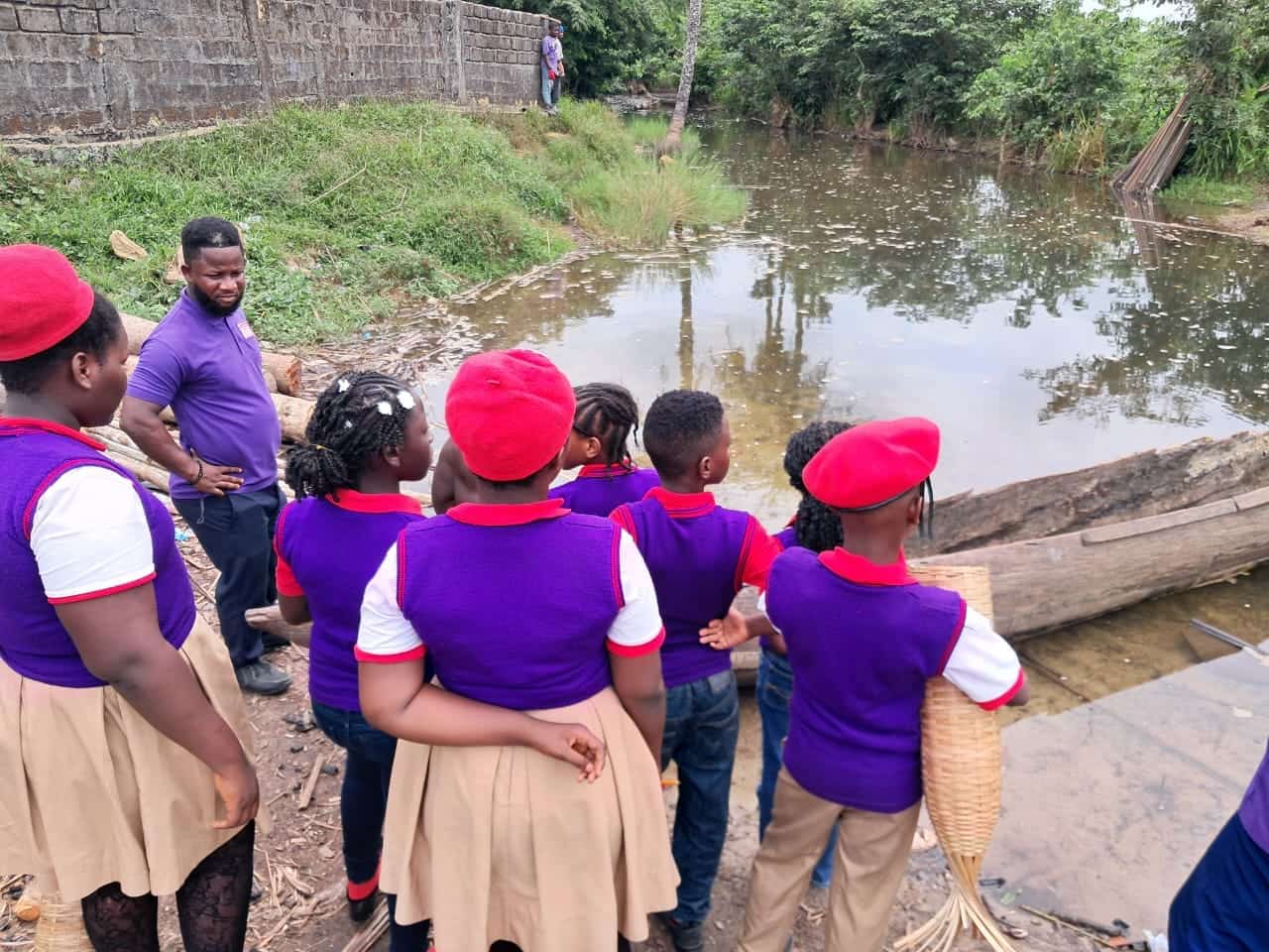 Children in red berets and purple shirts standing with a man beside a creek, one holding a woven basket.