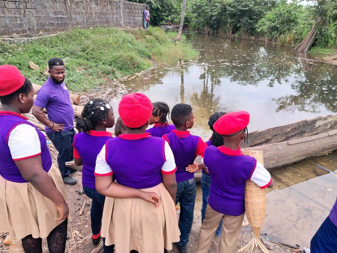 Children in red berets and purple shirts standing with a man beside a creek, one holding a woven basket.