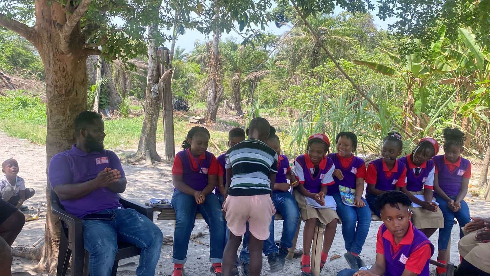 A man in a purple shirt sitting with a row of children in purple and red uniforms on benches under trees.
