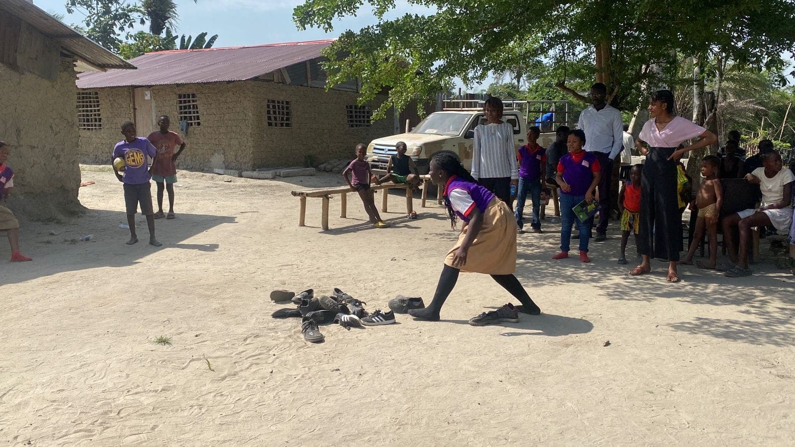 A girl bending over a pile of shoes during an outdoor game while children and adults watch from a sandy yard.