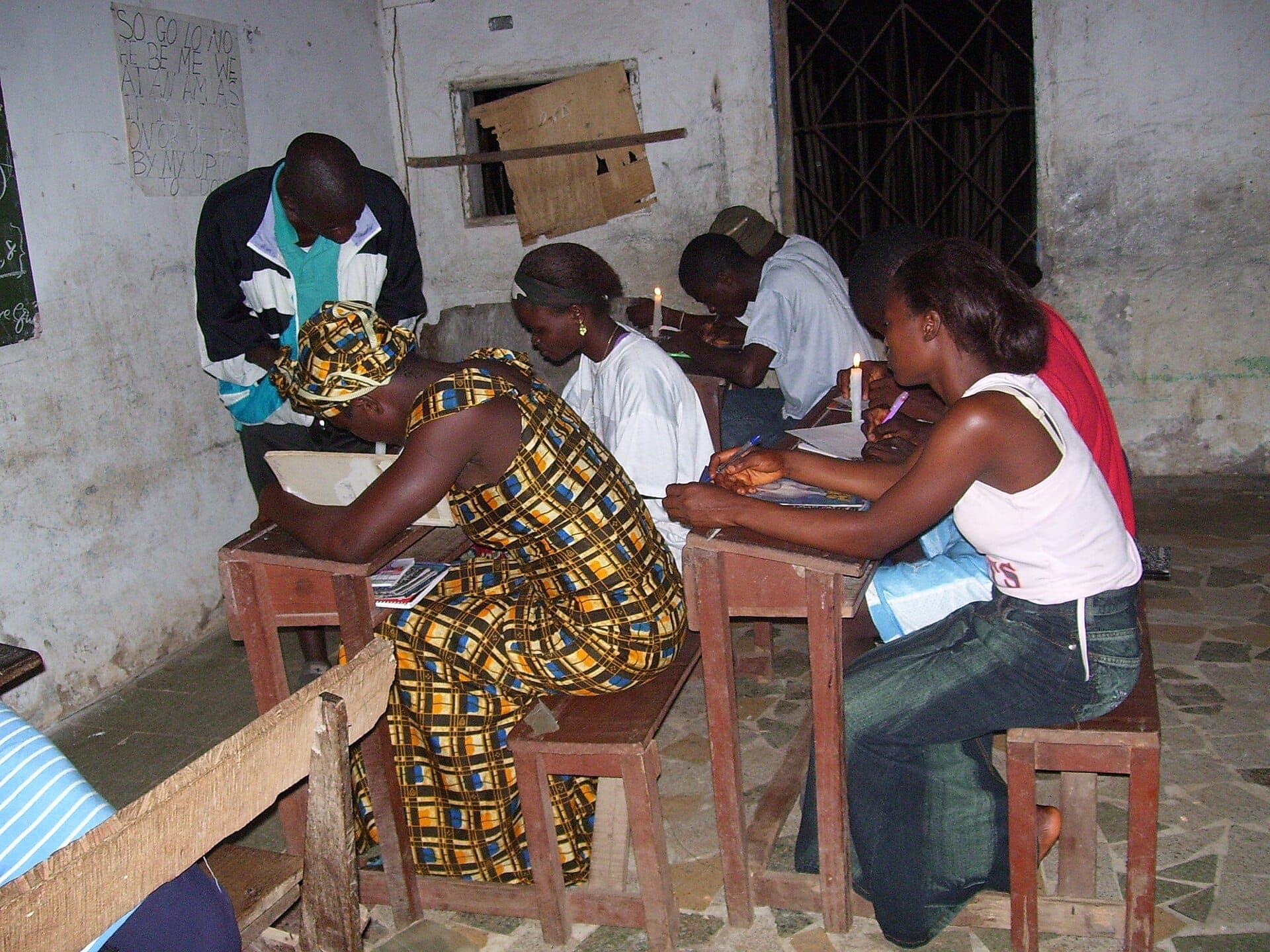 Students sitting at wooden desks writing in notebooks in a small classroom.